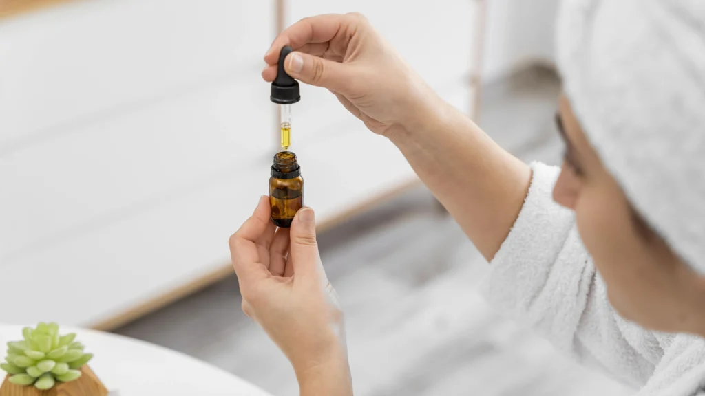 A woman in a white robe and towel turban incorporates a skincare routine by holding a small brown bottle with a dropper over her hand, preparing to apply the product