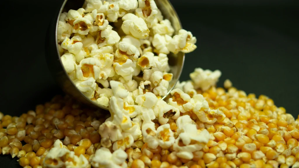 A bowl of freshly popped popcorn spilling onto a dark surface, surrounded by un-popped kernels. This scene captures the transformation from raw corn to fluffy popcorn, perfect for creating delicious snacks like nutritional yeast popcorn, which adds a savory and cheesy flavor without dairy 
