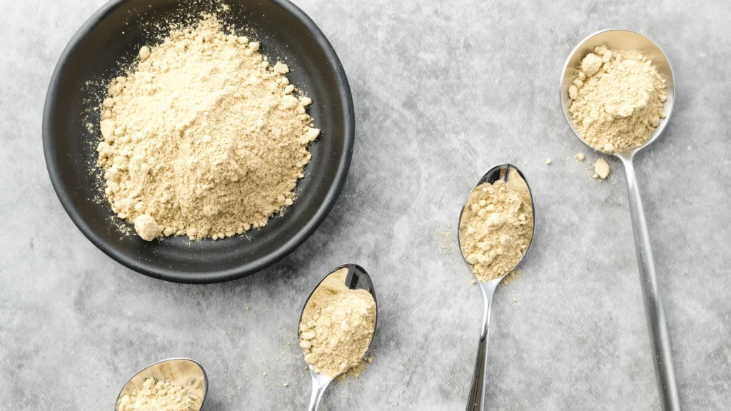 A bowl filled with a light beige powder, accompanied by three spoons each containing smaller portions of the same powder, arranged on a textured gray surface