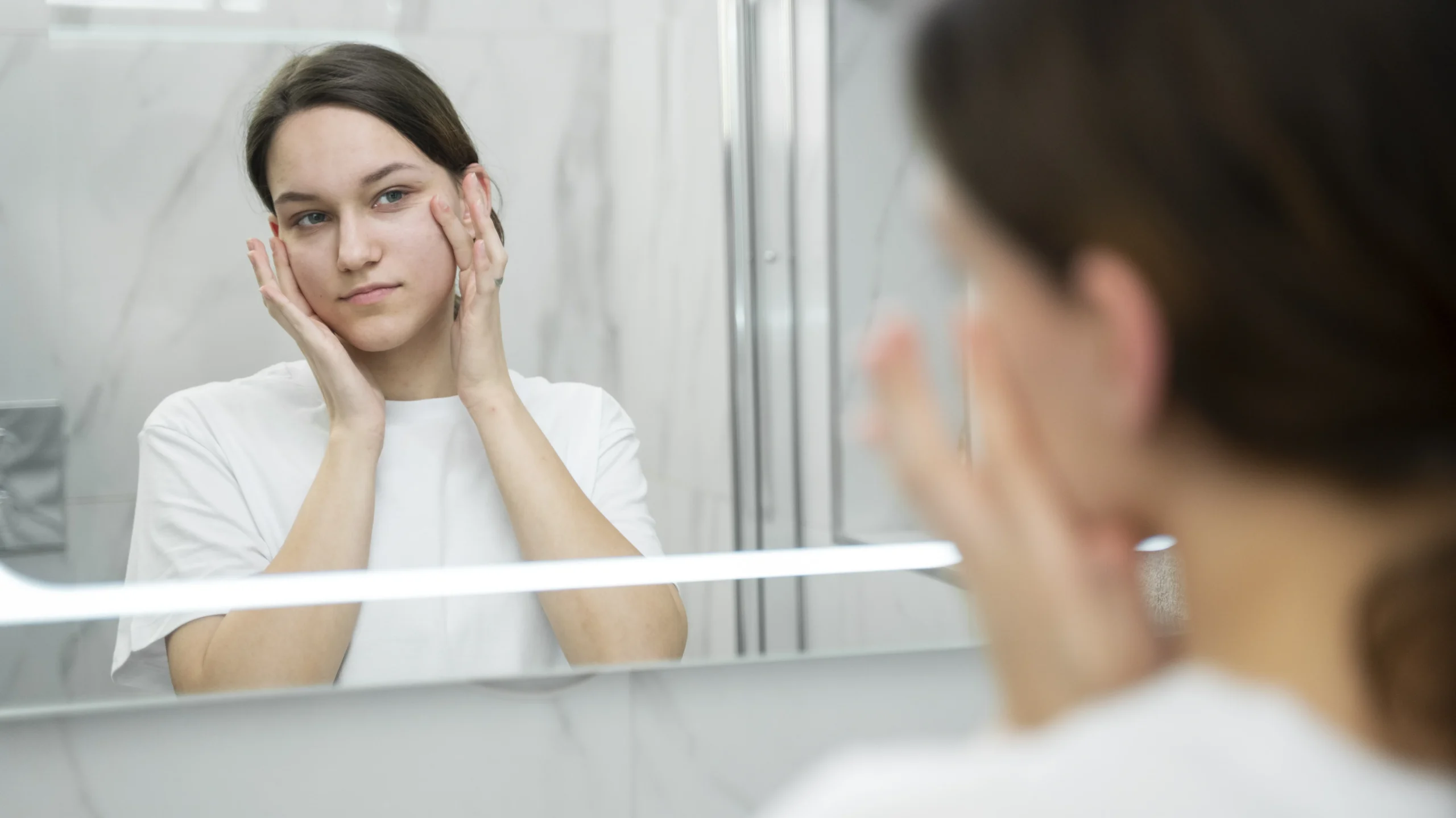 A woman in a white shirt is examining her face in the mirror, gently touching her cheek with both hands, highlighting the importance of understanding what causes dry skin.