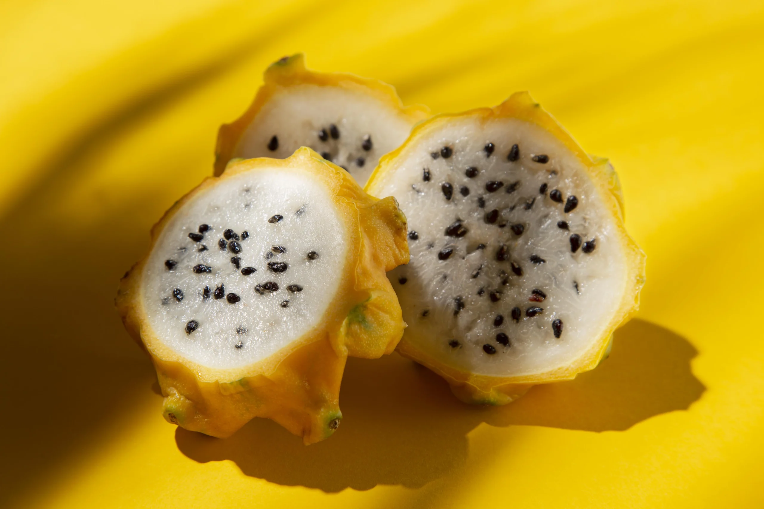 A close-up view of a sliced yellow dragon fruit, showcasing its creamy white flesh dotted with small black seeds. The vibrant yellow background highlights the fruit's unique texture and appearance.