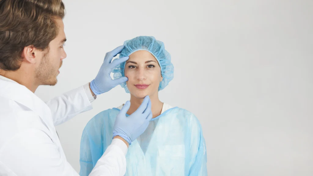 A doctor examines a patient's skin during a consultation. The patient is wearing a surgical cap and gown, while the medical professional checks for skin conditions. This image represents a scenario where someone might ask, What causes oily skin?