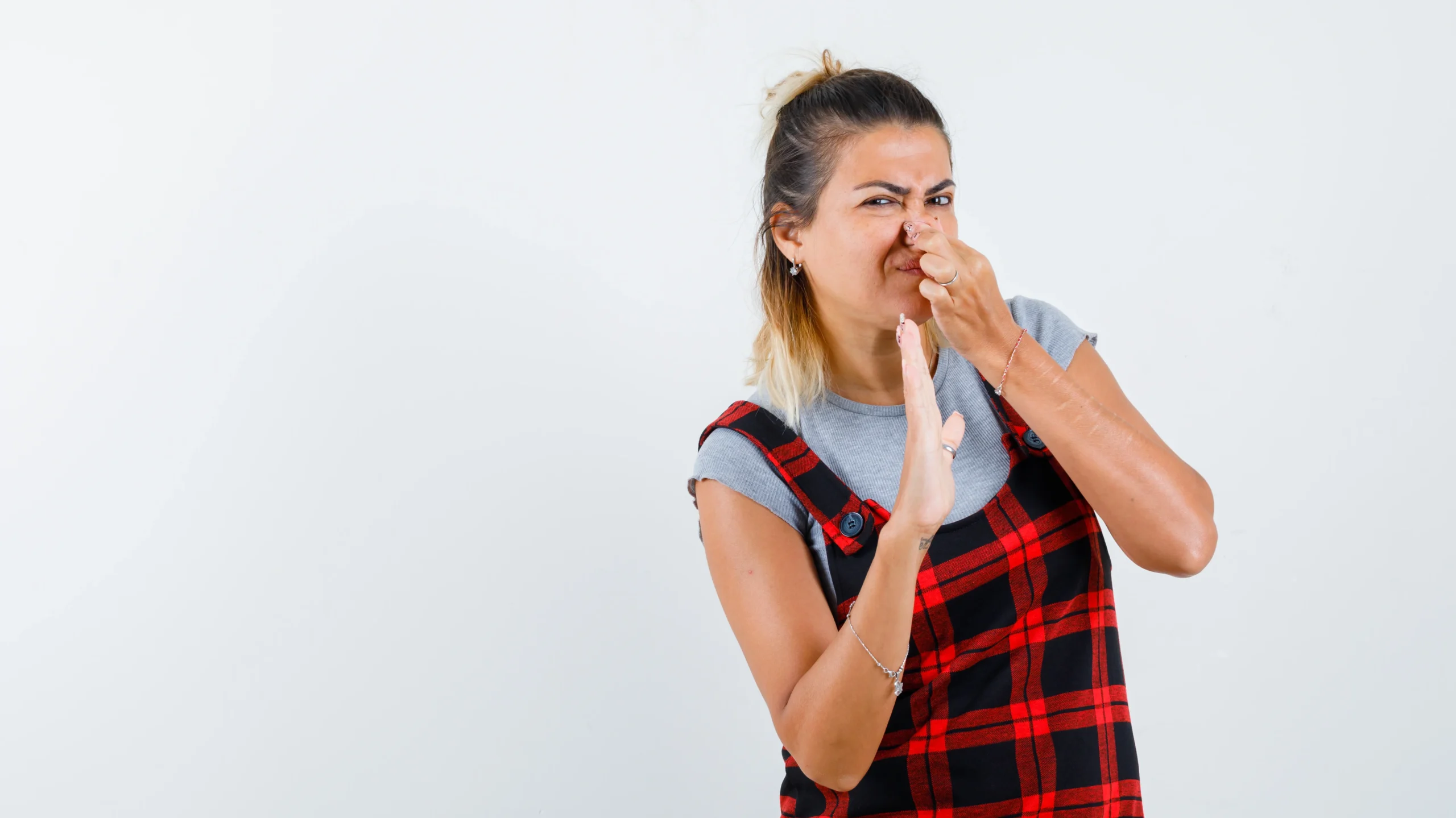 A girl wearing a red and black plaid overall over a gray shirt, holding her nose with one hand while making a disgusted expression. The image illustrates common reactions to unpleasant odors, such as those related to menstrual health concerns like Why Does My Period Smell