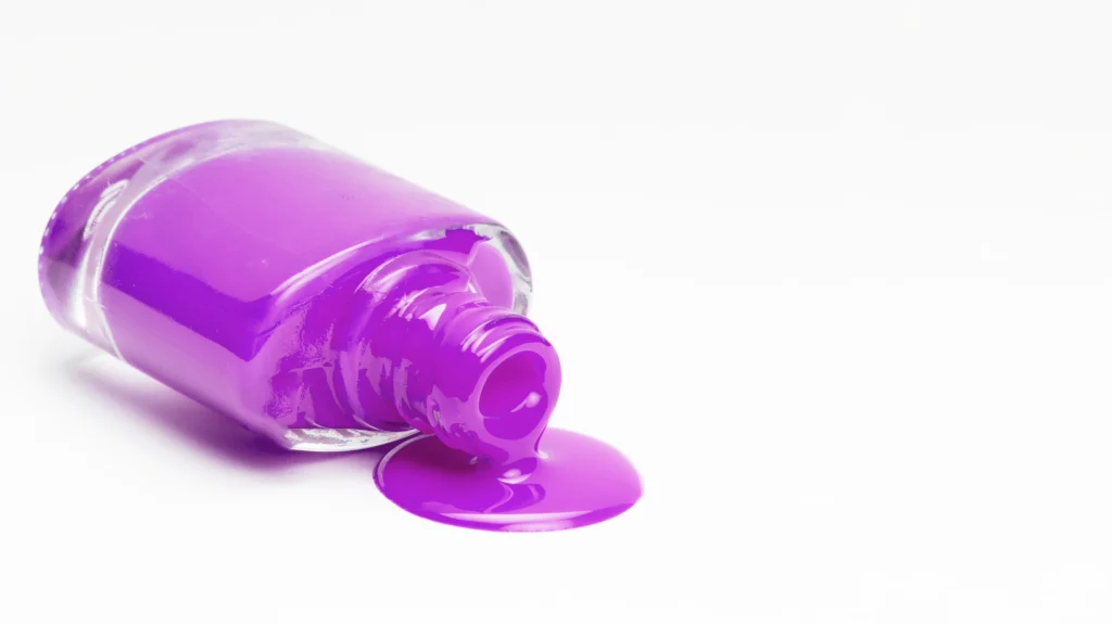 A vibrant purple bottle of nail polish lies on its side against a white background, with some of the bright, glossy polish spilling out.
