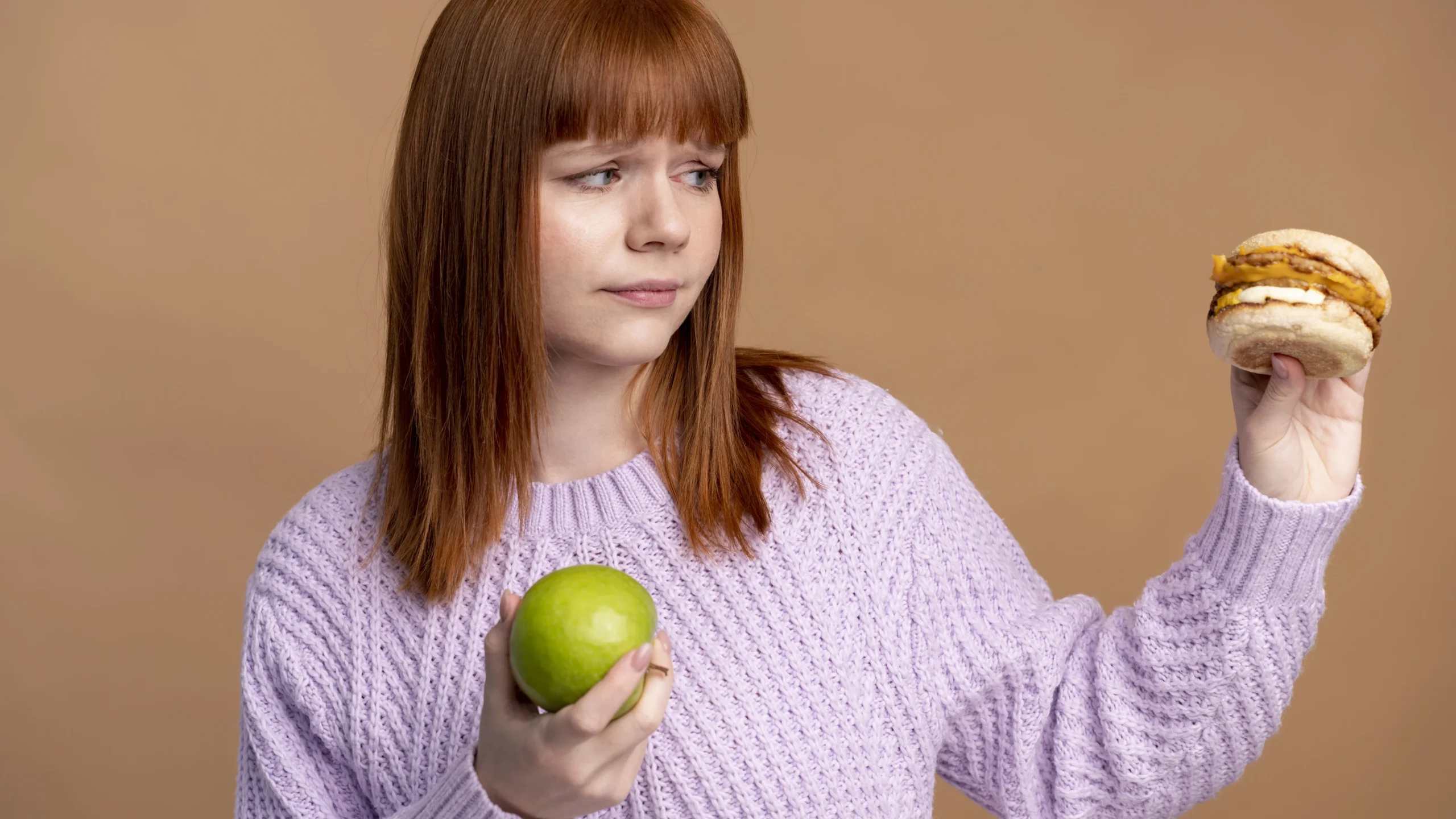 A woman with red hair and a purple sweater is holding a hamburger in one hand and a green apple in the other, symbolizing the importance of making mindful food choices to understand and address what causes dry skin.