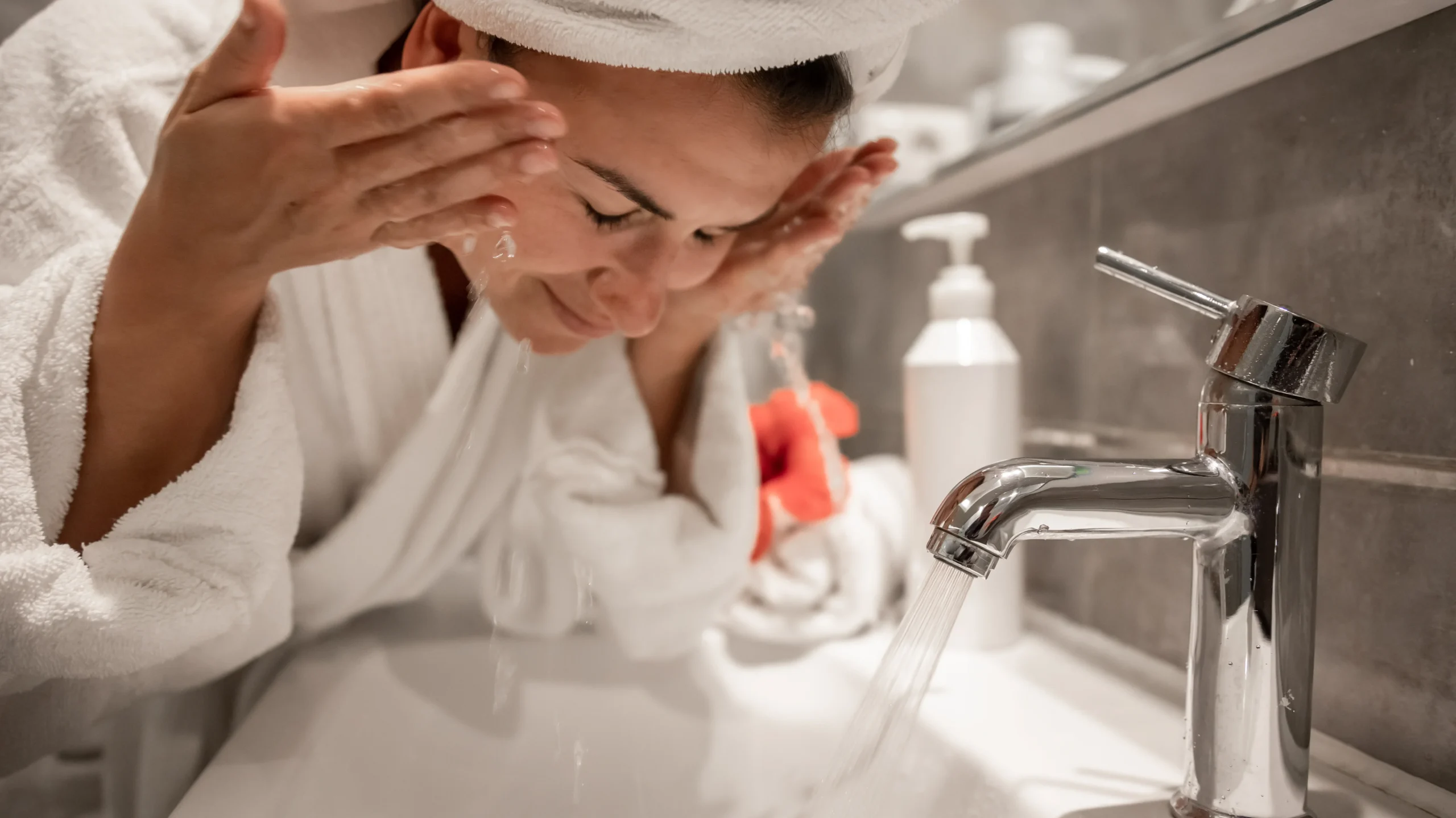 A woman with dark hair wrapped in a white towel is washing her face under running water at a sink, emphasizing the importance of understanding what causes oily skin.